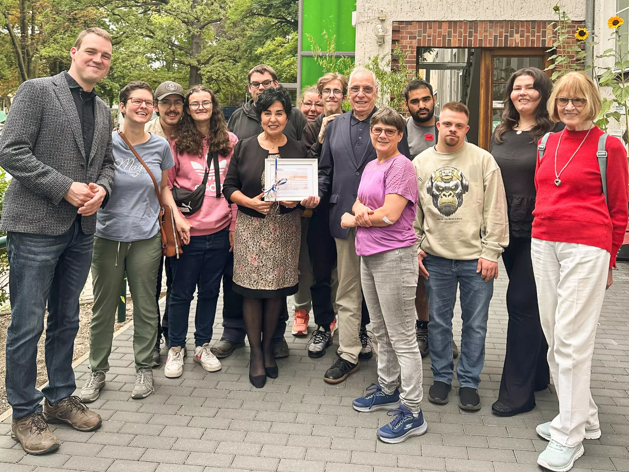 Gruppenfoto mit Bezirksbürgermeisterin Emine Demirbüken-Wegner und Spendencheck vor der Mosaik-Betriebsstätte in Reinickendorf. Außerdem abgebildet Betriebsstättenleiter Cornelius Haack sowie viele Beschäftigte und 2 Vertreter der Senioren