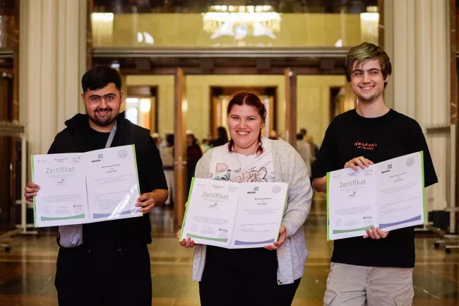 2 Absolventen und eine Absolventin von Mosaik halten stolz ihre Zertifikate im Roten Rathaus in die Kamera