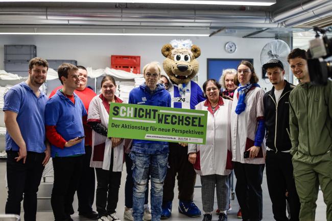 Auf dem Foto ist das Team aus der Wäscherei, zusammen mit Herthinho und 2 Hertha Spielern. Sie halten das Schild "Schichtwechsel" hoch.