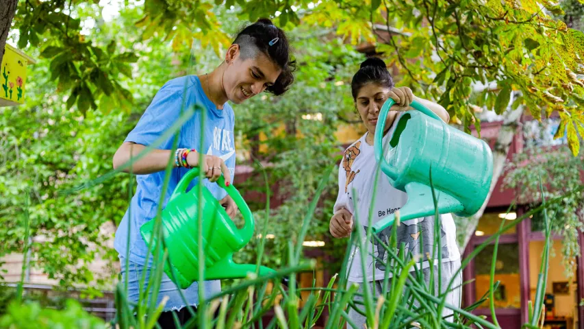 Gemeinsame Gartenarbeit im BFB Regina-Jonas-Straße - 2 Teilnehmerinnen gießen mit grünen Gießkannen die Hochbeete am Standort